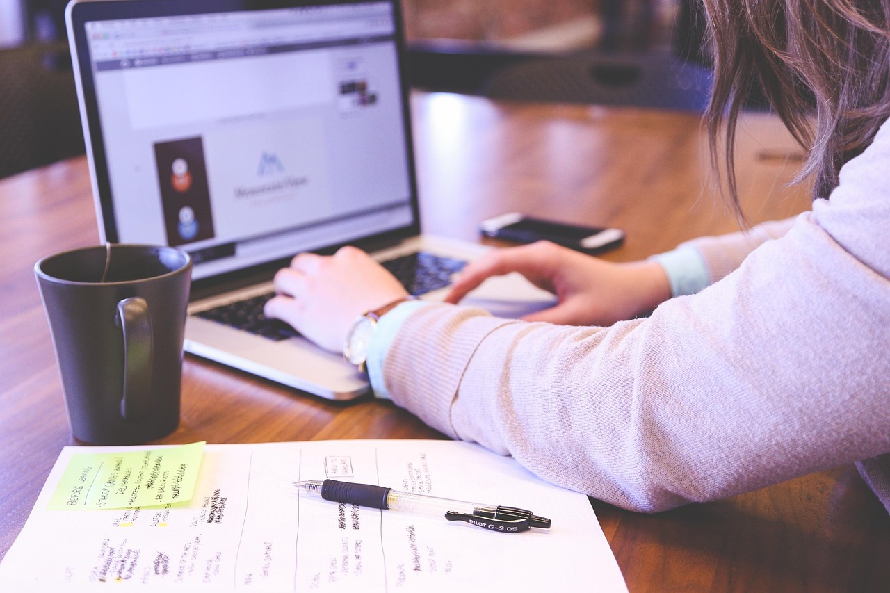 Support-Mitarbeiterin am Laptop mit handschriftlichen Notizen und Kaffeetasse am Schreibtisch